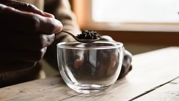 Hand pouring hot water over loose green tea leaves in a clear glass cup