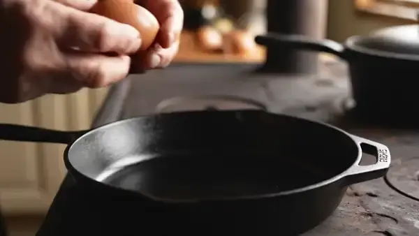 Hand cracking a brown egg into a black cast iron skillet on an old stove
